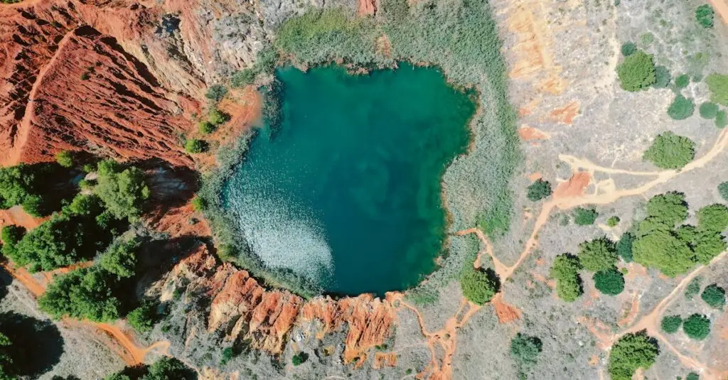 Drone view of turquoise water of small pond surrounded by rough stony formation with green plants